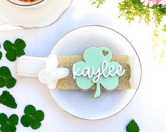 May include: A light green shamrock with a white heart and the name "kaylee" written in white cursive on a gold glitter background.