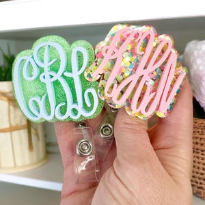 May include: Two glitter monogrammed badge holders with clear retractable badge reels. One holder is light blue with green glitter and the other is pink with multi-colored glitter.