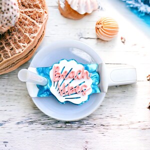 May include: A white seashell with the words "Beach Vibes" in pink lettering, attached to a blue and white patterned straw holder. The holder is attached to a white ceramic cup.