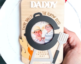 May include: A wooden Father's Day card with a photo frame shaped like a grill. The card says "Daddy, You're Grilly the Flippin' Best!" and "Happy Father's Day".