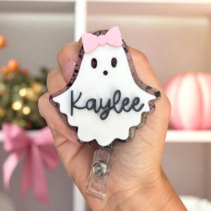May include: A white ghost-shaped badge reel with the name "Kaylee" in black script. The badge has a pink bow and a glittery border. The reel is held in a hand.