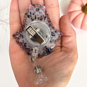 May include: A clear, ghost-shaped badge reel with iridescent glitter and a silver metal clip. The reel has a clear plastic strap and a retractable cord. A small pink pumpkin is in the background.