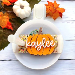 May include: A white ceramic mug with a handle and a pumpkin-shaped orange glitter decal with the name "kaylee" written in white cursive.