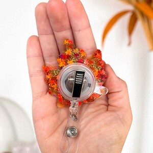 Puede incluir: Un portatarjetas con tem&aacute;tica oto&ntilde;al en forma de hoja de arce. La hoja est&aacute; llena de purpurina naranja, roja y dorada y peque&ntilde;as formas de hojas. El carrete tiene una carcasa de pl&aacute;stico transparente y un clip de metal negro.