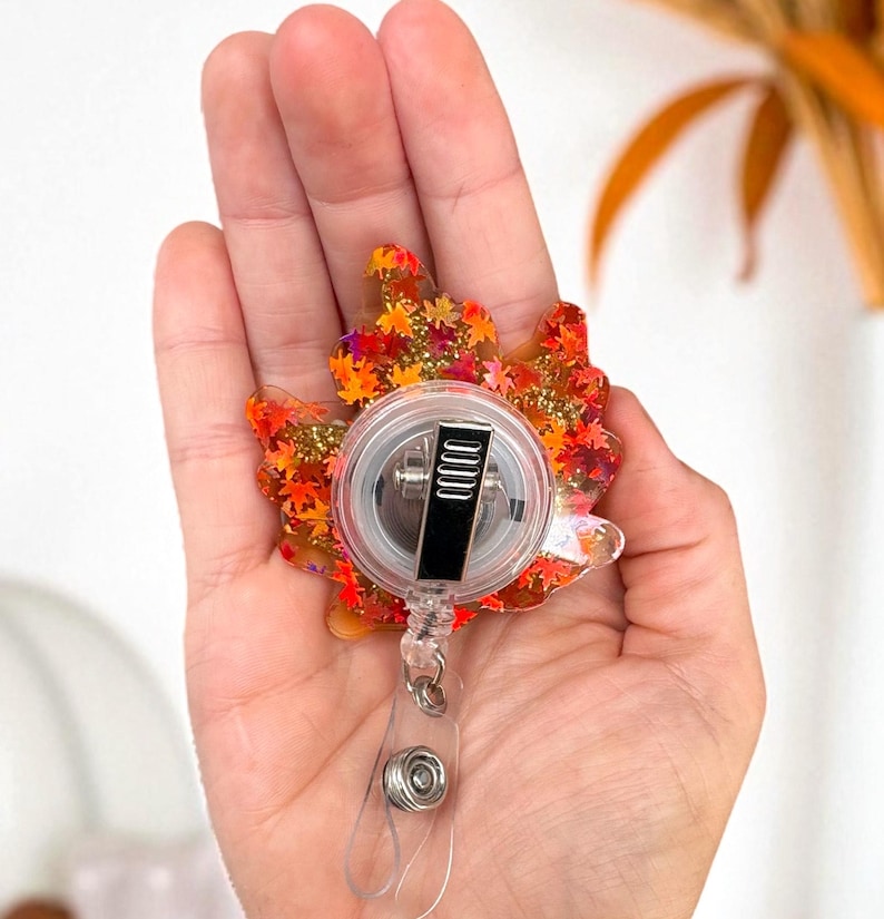 Puede incluir: Un portatarjetas con tem&aacute;tica oto&ntilde;al en forma de hoja de arce. La hoja est&aacute; llena de purpurina naranja, roja y dorada y peque&ntilde;as formas de hojas. El carrete tiene una carcasa de pl&aacute;stico transparente y un clip de metal negro.