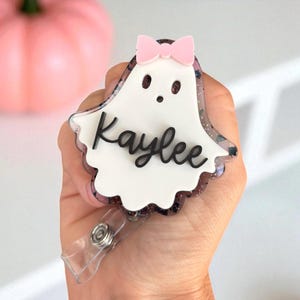 May include: A ghost-shaped badge reel with a pink bow and the name "Kaylee" in black script. The badge has a white center and a glittery, dark border. A clear plastic clip is attached to the bottom.
