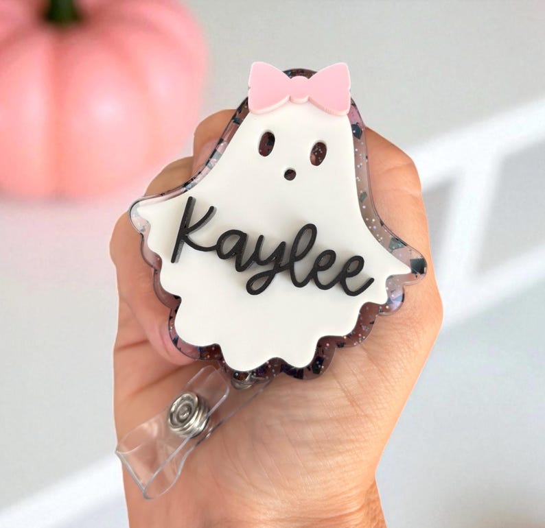 May include: A ghost-shaped badge reel with a pink bow and the name "Kaylee" in black script. The badge has a white center and a glittery, dark border. A clear plastic clip is attached to the bottom.