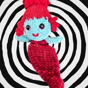 May include: A crocheted mermaid doll with a blue body, red hair, and a red tail. The doll is sitting on a black and white spiral background.