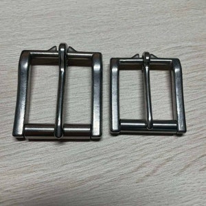 Może przedstawiać: Dwie srebrne metalowe klamry do paska, jedna większa od drugiej, na drewnianej powierzchni.