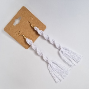 May include: White crocheted earrings with gold hooks and white tassels. The earrings are displayed on a brown card.