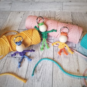May include: Three colorful crocheted keychain figures with wooden beads for heads. The figures are green, orange, and purple and are attached to key rings. The keychains are displayed on top of balls of yarn in pink, yellow, and white.