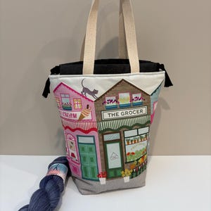 May include: A fabric tote bag featuring a colourful illustration of shops, including an ice cream shop and a grocer. The bag has cream-coloured handles and a dark grey interior. A skein of blue yarn rests beside the bag.