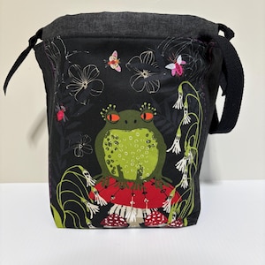 May include: A black drawstring bucket bag featuring a graphic print of a green frog with orange eyes sitting on a red mushroom. The bag includes a dark grey top band, a black shoulder strap, and a botanical pattern with white flowers and insects.