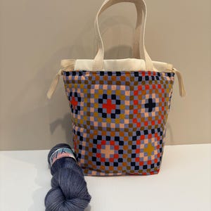 Peut inclure: Un sac fourre-tout coloré avec un motif géométrique dans les tons de bleu, rouge, orange et marron. Le sac a un intérieur et des poignées de couleur crème. Une pelote de laine bleue est à côté du sac, suggérant un thème de tricot ou d'artisanat.