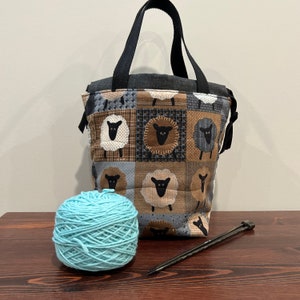 May include: A black and brown tote bag with a patchwork design featuring sheep. The bag has a black handle and is made of fabric. A ball of light blue yarn and a pair of knitting needles are also in the image.