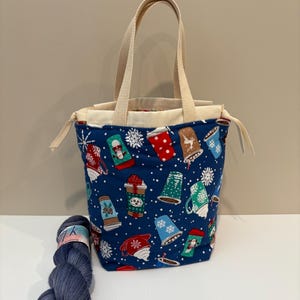 May include: A blue tote bag with cream-coloured interior and handles. The bag features a pattern of coffee cups and snowflakes. A skein of blue yarn is placed beside the bag.
