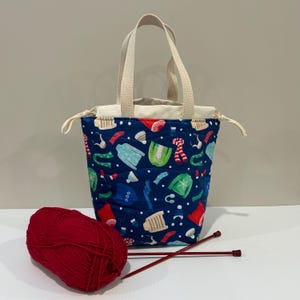 May include: A blue canvas tote bag with a drawstring closure and white handles. The bag is decorated with a pattern of colourful winter clothing, including sweaters, scarves, hats, and mittens. A ball of red yarn and two red knitting needles are in front of the bag.