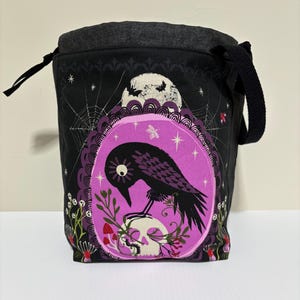 Peut inclure: Un sac à cordon noir avec un cadre ovale violet central contenant un corbeau noir perché sur un crâne. Le motif comprend des toiles d'araignée blanches, une lune avec des chauves-souris, des petits champignons rouges et du feuillage vert.