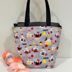 May include: A grey tote bag with black handles, decorated with a repeating pattern of cartoon chickens. A skein of pink and orange yarn sits in front of the bag, suggesting a knitting or crafting theme.