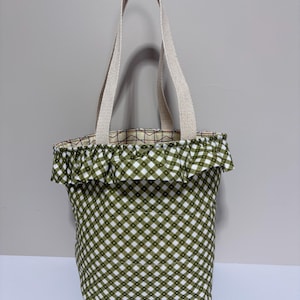 May include: A tote bag featuring a green and white gingham pattern with a ruffled top edge. The bag has two beige woven fabric handles. The interior lining is visible at the top opening, displaying a geometric grid pattern in red, black, and white.