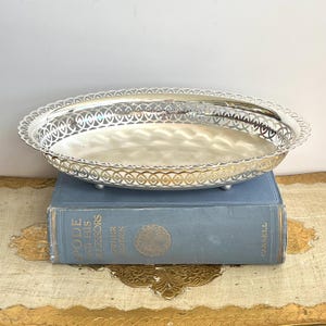 Peut inclure: Un plateau de service ovale de couleur argentée doté d'une bordure ajourée décorative et d'une base lisse et réfléchissante. Le plateau est soutenu par de petits pieds et repose sur un livre à couverture bleue intitulé Spode and His Successors.