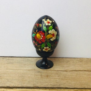 Peut inclure: Un œuf en bois noir avec un motif floral peint à la main dans des tons de rouge, jaune, blanc et vert. L'œuf est présenté sur un support en bois noir.