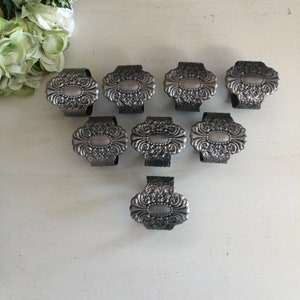 May include: Set of eight silver napkin rings with an ornate design. Each ring features a raised oval with a textured surface and intricate floral details.