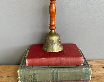 May include: A brass bell with a wooden handle. The bell has a floral design and is sitting on top of a stack of three books.