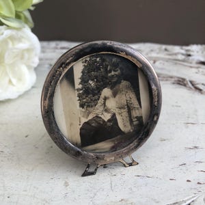 Peut inclure: Un cadre photo rond vintage, de couleur argentée, avec une photographie en noir et blanc. Le cadre a une finition vieillie et un petit support. La photographie représente une femme en haut en dentelle.