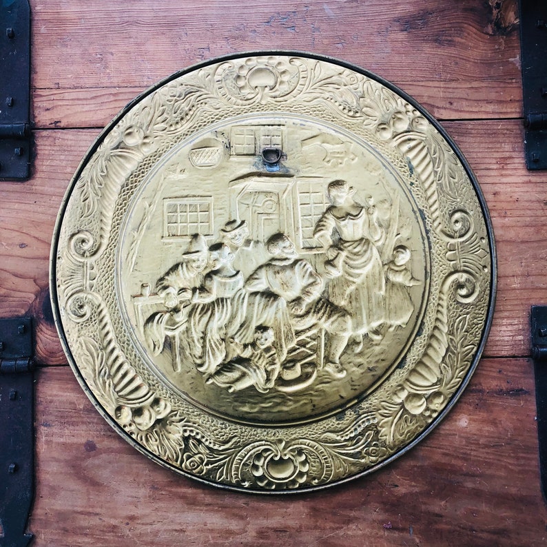 May include: A round, brass-colored decorative plate with a detailed relief scene of people gathered outside a building. The plate has an ornate floral border and is mounted on a wooden surface. The relief depicts figures in period clothing.