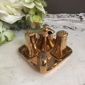 May include: Gold-toned ceramic salt and pepper shakers and a condiment dish with a lid, all on a matching square tray.