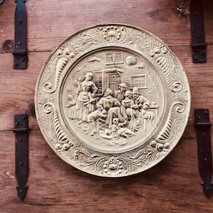 May include: A round, gold-colored decorative plate with a detailed relief scene of people gathered outside a building. The plate has an ornate, floral border and is mounted on a wooden surface with black hinges.
