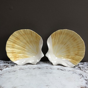 May include: Two ceramic seashell-shaped dishes with a white interior and a yellow and brown exterior.