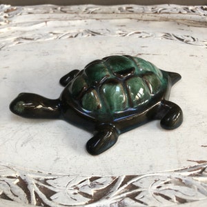 May include: A ceramic turtle figurine with a dark green and brown glaze. The turtle's shell has a detailed design and the figurine is sitting on a white surface.