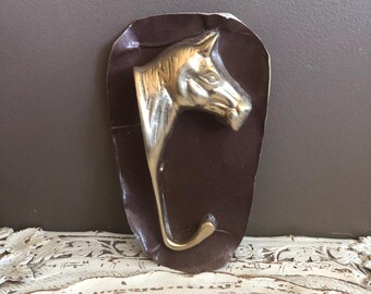 May include: A brass horse head hook mounted on a dark brown, irregularly shaped backing. The hook is a stylized horse head with a curved hook extending from the neck. The piece is designed for hanging items.