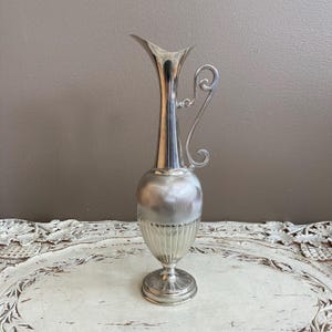 May include: A tall, silver-colored vase with a flared top and a decorative handle. The vase has a bulbous midsection and a fluted base. The overall design is elegant and vintage, suitable for floral arrangements or display.