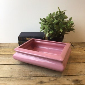 May include: A pink ceramic planter with a rectangular shape and a slightly raised rim. The planter is sitting on a wooden surface.