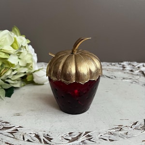 May include: A ruby red glass strawberry-shaped jar with a gold-toned metal lid and stem. The jar has a textured surface. The lid is intricately detailed. The jar is set against a neutral background, evoking a sense of vintage charm and elegance.