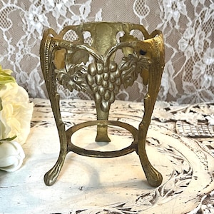 May include: A vintage brass egg holder with a grape and leaf design. The holder has a three-legged base and a circular support ring. The top features an openwork design. The brass has a warm, aged patina, suggesting it is an antique or vintage piece.
