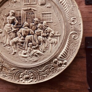 May include: A decorative brass plate with a detailed relief scene of people gathered outside a building. The plate has a detailed floral and scrollwork border. The scene depicts figures in period clothing, suggesting a historical or vintage theme.