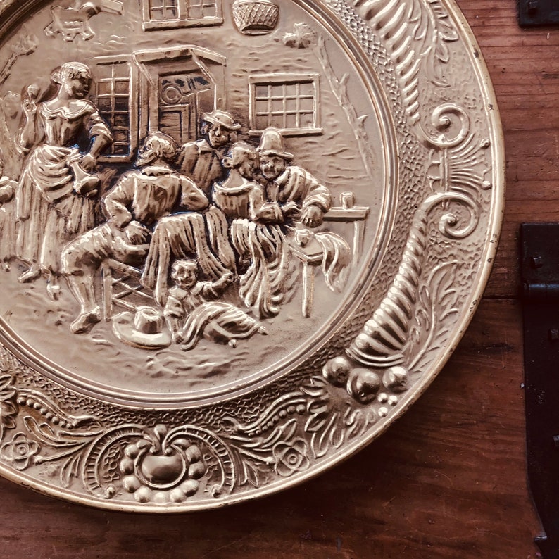 May include: A decorative brass plate with a detailed relief scene of people gathered outside a building. The plate has a detailed floral and scrollwork border. The scene depicts figures in period clothing, suggesting a historical or vintage theme.