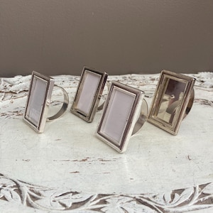 May include: Four silver-toned rectangular napkin rings with a picture frame design. Each ring has a rectangular frame with a light-colored insert and a curved band for the napkin. The rings are displayed on a white, distressed surface.