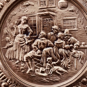 May include: A silver-toned decorative plate with a detailed relief scene of people gathered outside a building. The relief depicts figures in period clothing, with architectural details like windows and a basket.