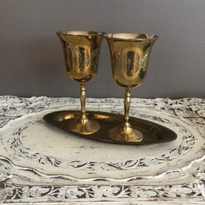 May include: Two gold-tone metal goblets with ornate designs on a gold-tone metal tray. The tray has a beaded edge.
