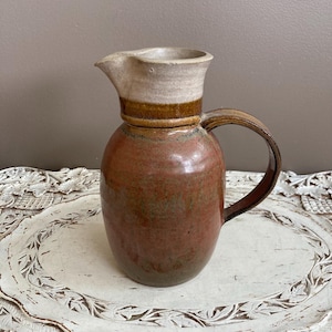May include: A handmade ceramic jug with a rounded body and a spout. The jug features a reddish-brown base, a tan rim, and a brown handle. The jug is sitting on a white, carved tray.