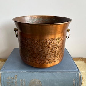 Puede incluir: Un cubo de cobre con una abertura ancha y un patrón texturizado de estilo martillado en el exterior. El recipiente cuenta con dos pequeños anillos de metal de color latón unidos a los lados opuestos cerca del borde.