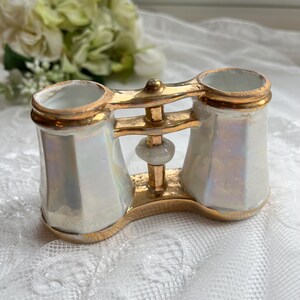 May include: A vintage, iridescent white porcelain decorative piece, shaped like opera glasses, with gold-coloured accents. The piece has two cylindrical openings and a connecting gold-coloured frame. The base is also gold-coloured.