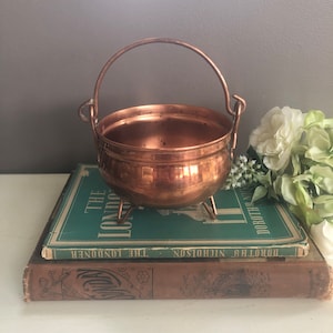 May include: A small copper pot with a handle, sitting on top of two books. The pot has a hammered finish and three legs. The books are green and brown.