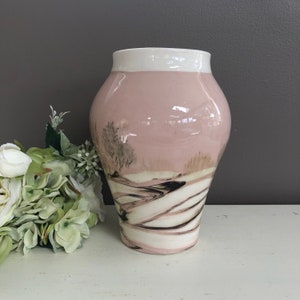 May include: A pink ceramic vase with a white rim and a black and white landscape design. The vase has a rounded body and a narrow neck.