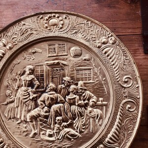 May include: A brass-colored decorative plate with a detailed relief scene of people gathered outside a building. The plate has an ornate floral border and a central scene depicting figures in period clothing. The plate is displayed on a wooden surface.